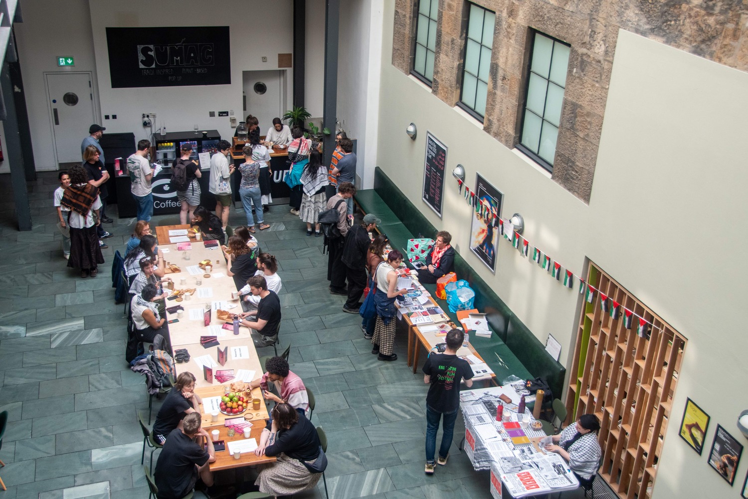 Photo taken from above of people sitting & talking, queuing for food and using the book exchange at Falastin Film Festival.