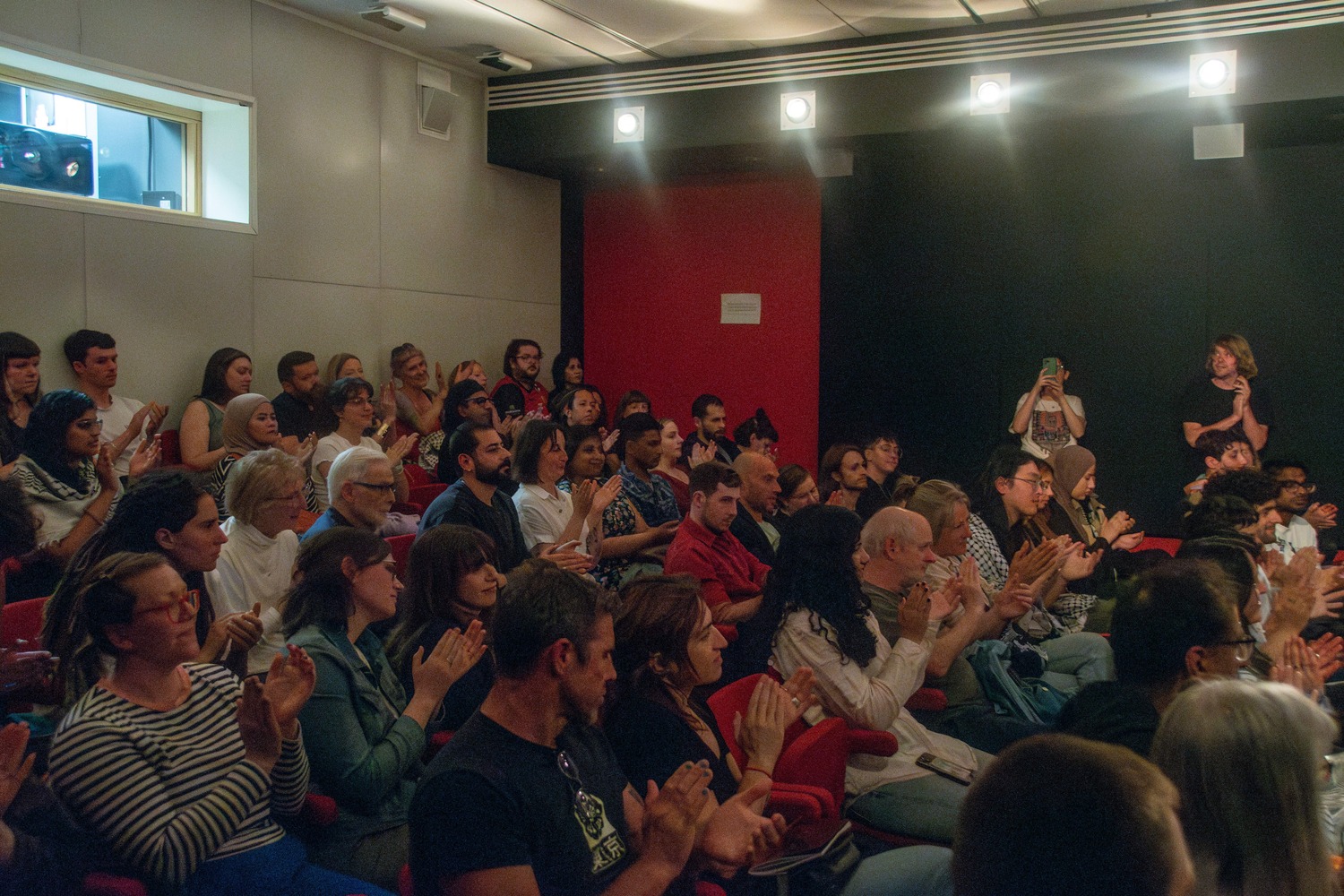 An audience at Falastin Film Festival sitting in red cinema chairs and applauding at the end of one of the films shown.
