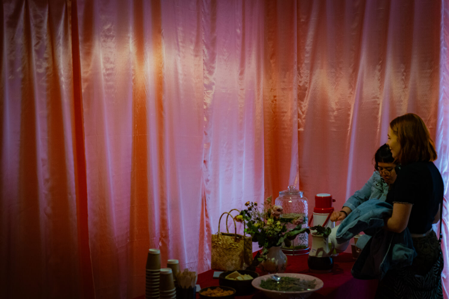 The table set up with tea and water in the Listen Gallery. The walls are draped in pink silk. Two people chat and pour themselves tea.